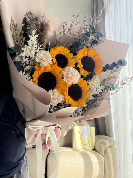 Bouquet of sunflowers and white flowers in pink paper wrap held by a person.