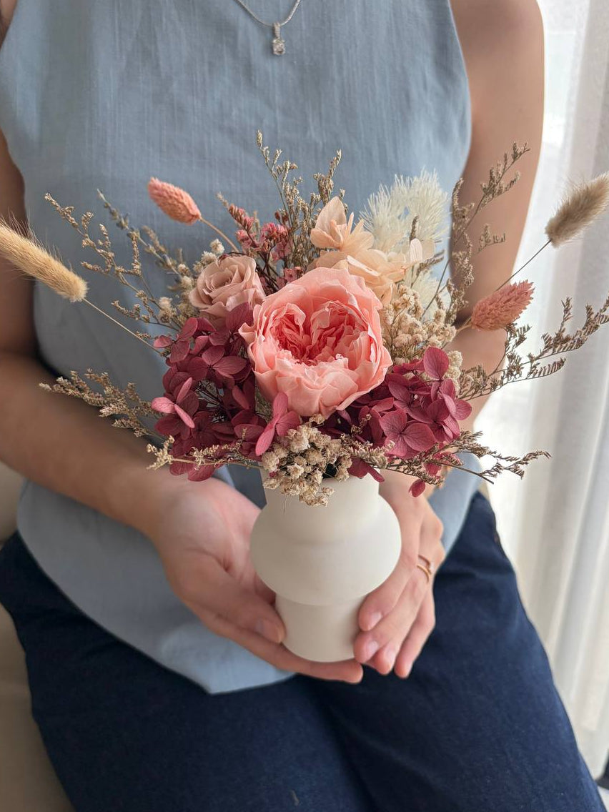 Person holding a small white vase with a bouquet of pink and beige flowers.