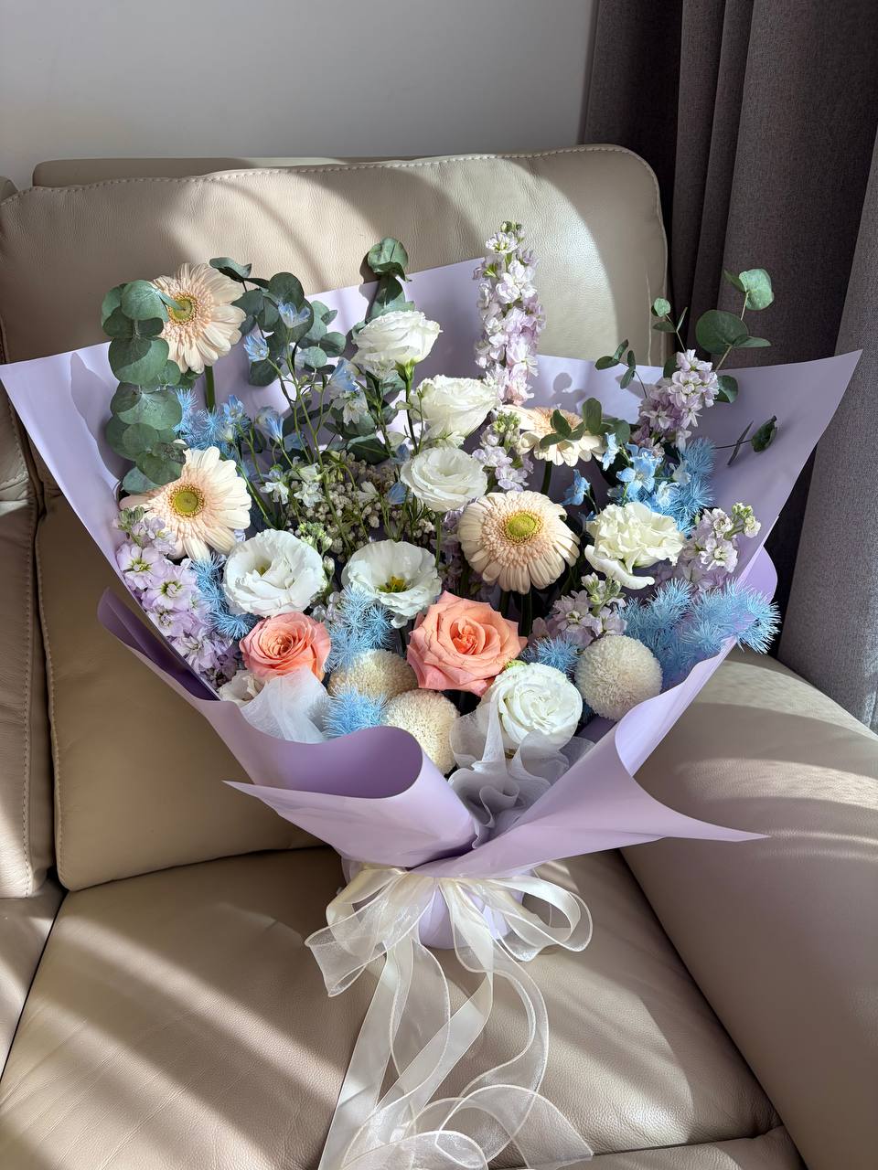 A large, beautifully arranged floral bouquet featuring white and pink roses, daisies, eucalyptus, and pastel purple flowers, wrapped in light purple paper and tied with a sheer white ribbon, resting on a beige leather armchair.