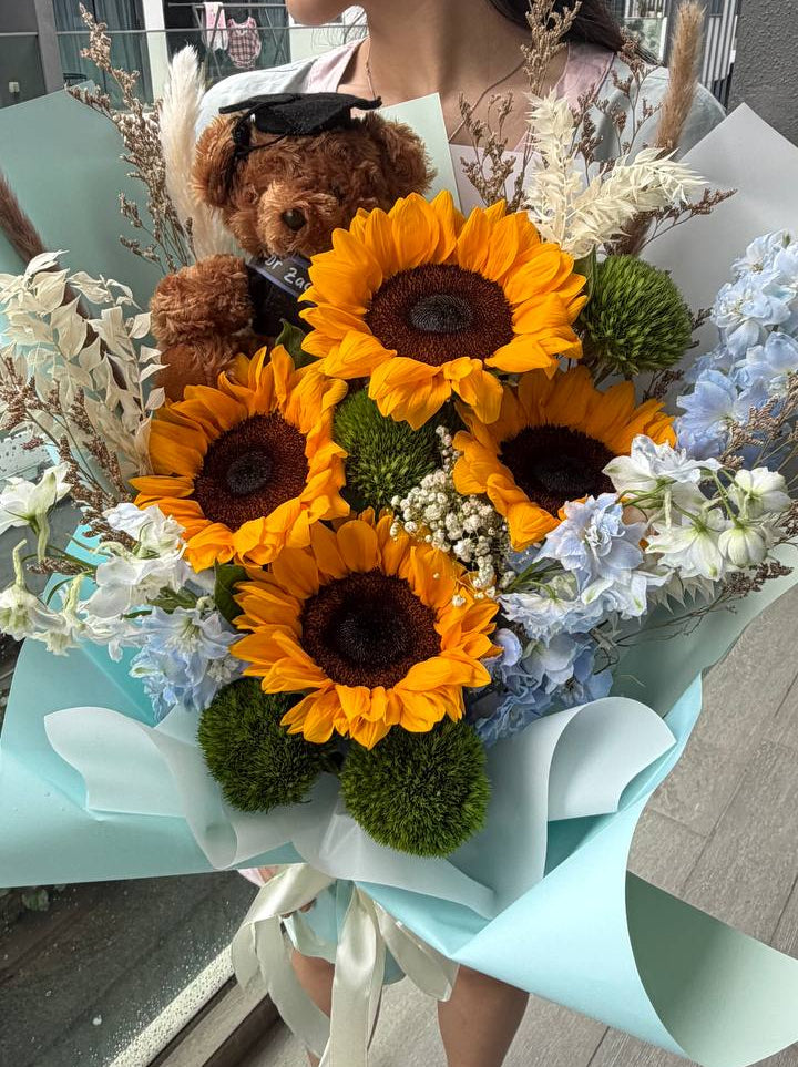 A person holding a large sunflower bouquet with a small brown teddy bear tucked inside, wrapped in pale blue paper. The bouquet includes bright yellow sunflowers, white and blue flowers, green foliage, and dried grasses, photographed in an indoor setting.