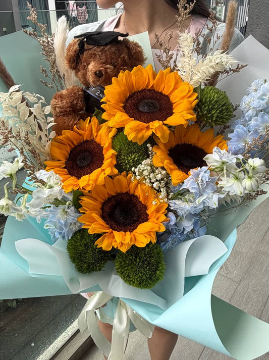 A person holding a large sunflower bouquet with a small brown teddy bear tucked inside, wrapped in pale blue paper. The bouquet includes bright yellow sunflowers, white and blue flowers, green foliage, and dried grasses, photographed in an indoor setting.