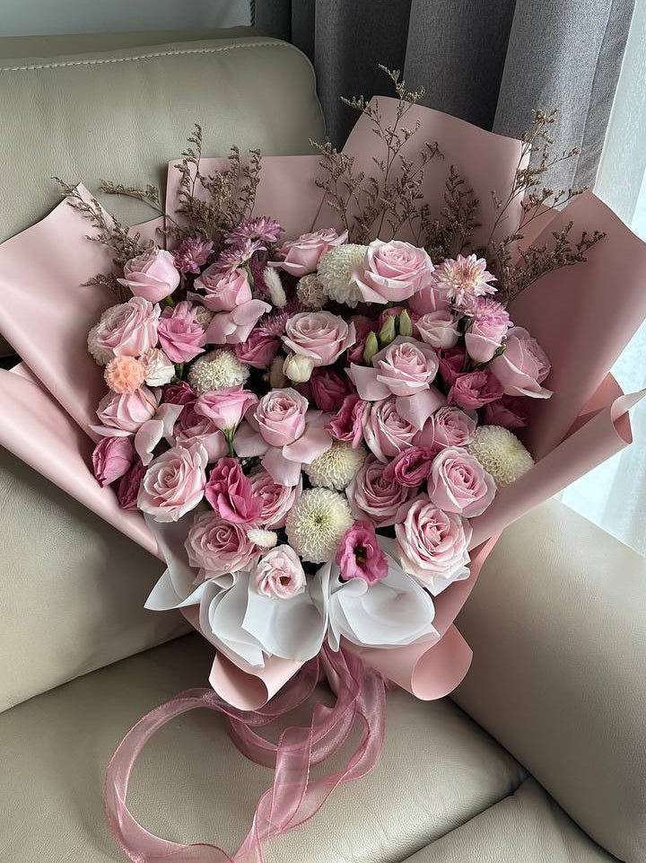 A bouquet of pink and light pink roses wrapped in pink paper, resting on a beige leather sofa in front of gray curtains.