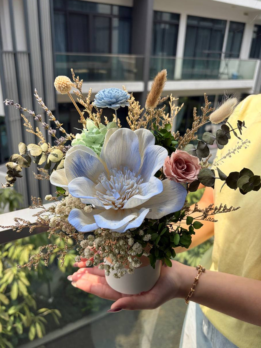 A person holding a decorative dried flower arrangement in a white pot, featuring a large pale blue flower, a pink rose, green foliage, and beige filler stems, with a modern building in the background.