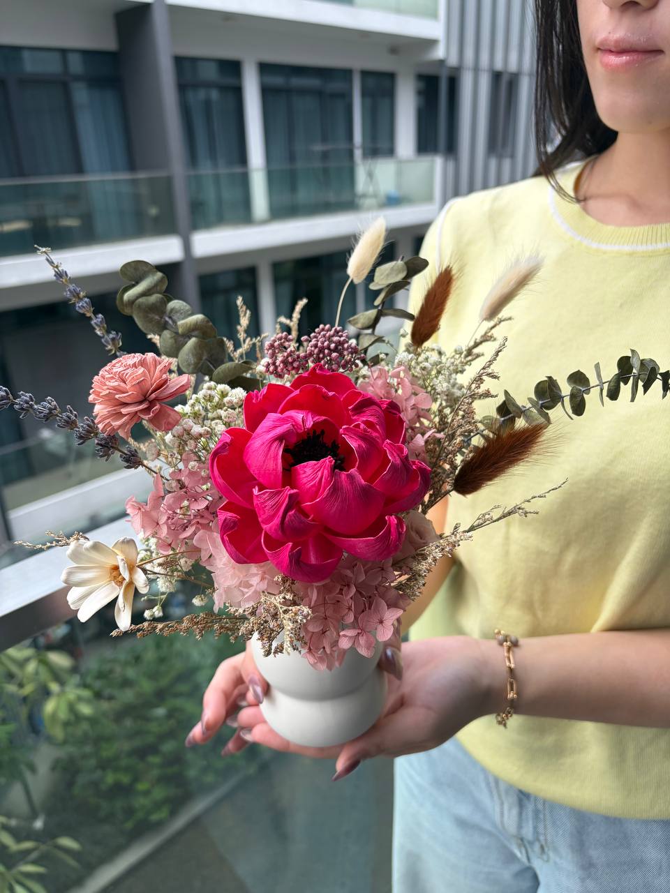 A woman in a yellow top holds a dried flower arrangement in a white vase on a balcony. The arrangement features a prominent bright pink peony, a dusty pink rose, a white daisy, and green eucalyptus stems.