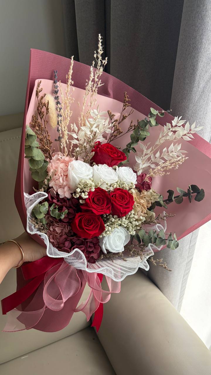 A dried flower bouquet wrapped in pink paper with a pink ribbon, held by a hand near a beige sofa and gray curtains.